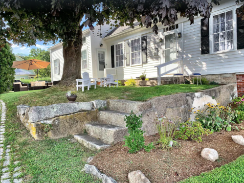 A charming house with a stone pathway, garden, and outdoor seating under a large tree and an umbrella.