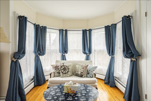 Cozy living room with a white sofa, nautical-themed pillows, and blue curtains framing large windows.