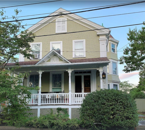 A large yellow house with a porch, surrounded by greenery and power lines, under a clear blue sky.