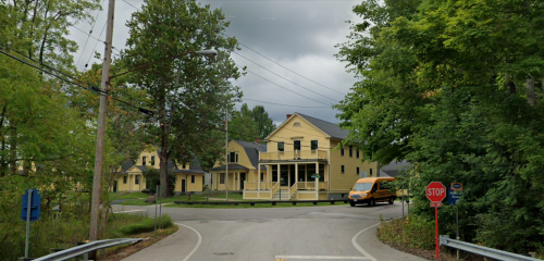 A yellow house at a road intersection, surrounded by trees, with a stop sign and a van approaching. Cloudy sky above.