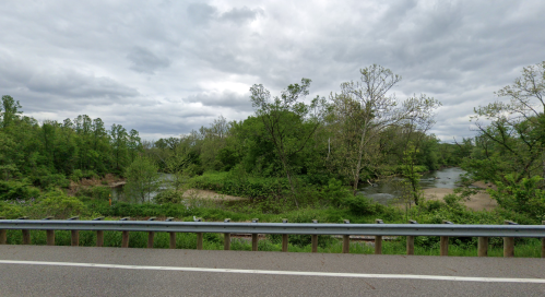 A scenic view of a lush green landscape with a river, under a cloudy sky, alongside a road with a guardrail.