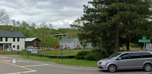 A construction site with new homes, a gray van on the road, and trees under a cloudy sky.