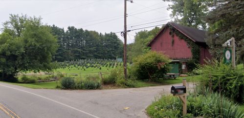 A red barn near a road, surrounded by greenery and a field of trees in the background under a cloudy sky.