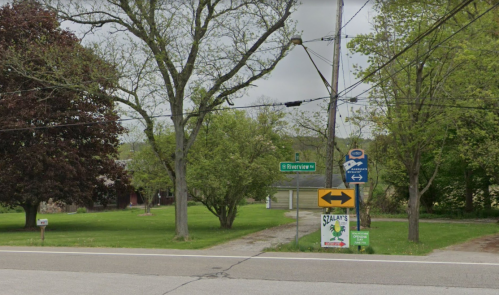 A street view showing a green sign for E Riverine, trees, and a directional sign near a grassy area.