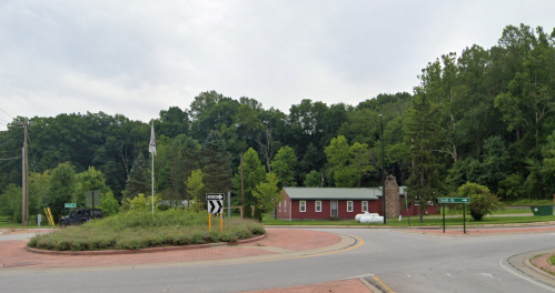 A roundabout with a red building and greenery in the background, surrounded by trees and a cloudy sky.