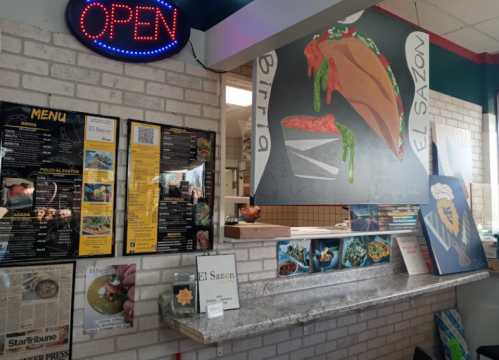 Interior of a restaurant featuring a menu board, an "Open" sign, and colorful artwork of tacos and birria.