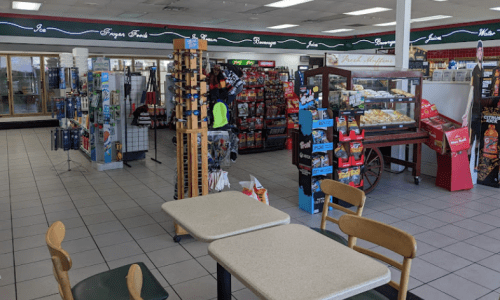 Interior of a convenience store with shelves of snacks, drinks, and a seating area with two tables.