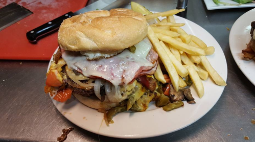 A large burger topped with cheese, ham, and vegetables, served with a side of crispy French fries on a white plate.