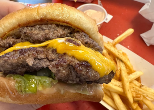 A hand holding a cheeseburger with lettuce and a side of fries on a red table.