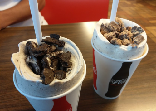Two cups of ice cream topped with crushed cookies, one with chocolate and the other with a mix of toppings, on a table.