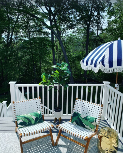 Two chairs with tropical cushions on a deck, surrounded by greenery, with a striped umbrella and a potted plant nearby.