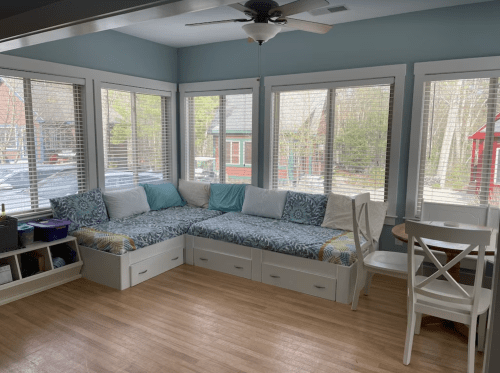 Bright sunroom with a cozy corner seating area, large windows, and a small dining table. Light wood flooring.