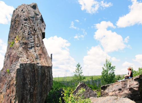 A person sits on a rock, gazing at a large boulder under a blue sky with fluffy clouds and greenery around.