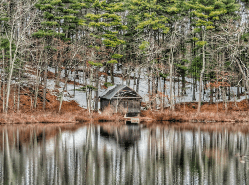 A rustic cabin by a calm lake, surrounded by trees and a hint of snow on the ground. Reflections in the water.