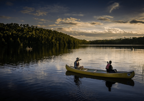 Two people in a yellow canoe on a calm lake at sunset, surrounded by trees and distant kayakers.