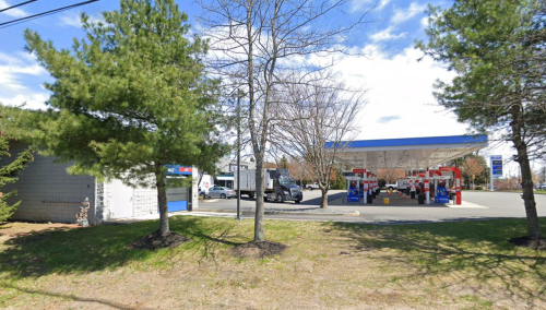A gas station with a blue roof, surrounded by trees and a clear sky, with vehicles parked nearby.