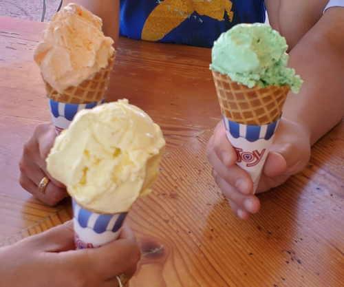 Three hands hold ice cream cones with orange, green, and yellow scoops against a wooden table background.