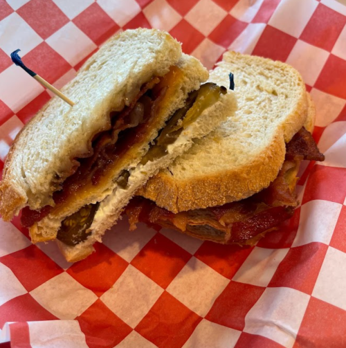 A sandwich with layers of bacon, cheese, and pickles, served on sliced bread with a toothpick, on a red and white checkered paper.