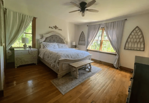 Cozy bedroom with a large bed, wooden floors, and natural light from windows, featuring elegant decor and soft curtains.