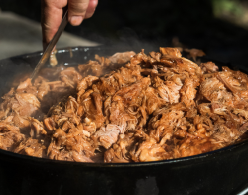A close-up of tender, shredded meat being stirred in a large pot, with steam rising in the background.