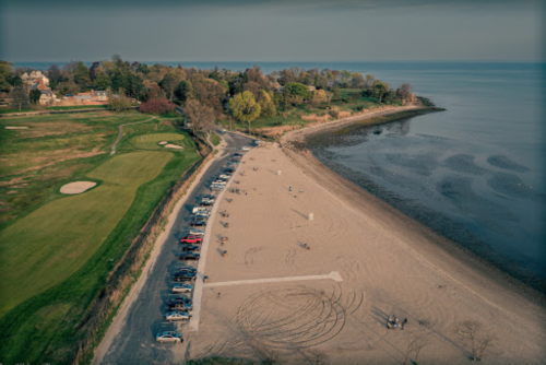 Aerial view of a beach with parked cars, a golf course, and trees along the shoreline under a clear sky.