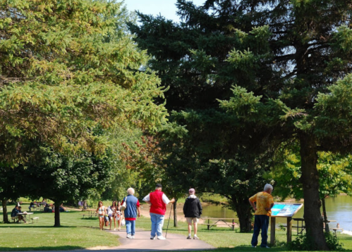A sunny park scene with people walking along a path, surrounded by trees and a river in the background.