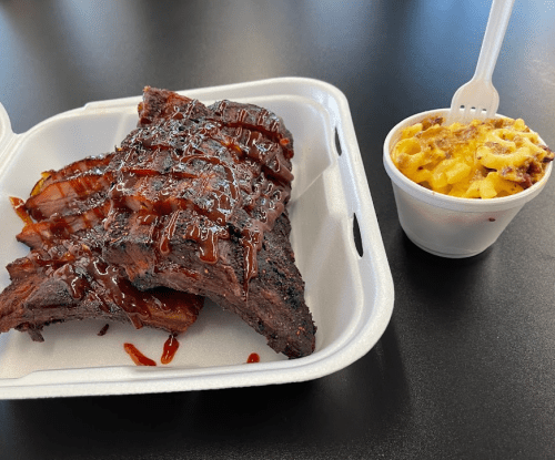 A takeout container with BBQ ribs drizzled in sauce, alongside a cup of macaroni and cheese.
