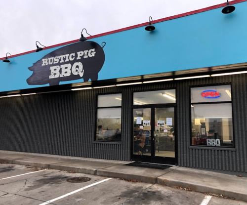 Exterior of Rustic Pig BBQ restaurant, featuring a blue sign and open door, with parked cars in front.