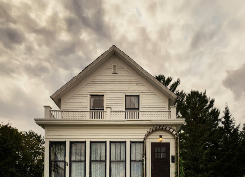 A white two-story house with a dark door and large windows, set against a cloudy sky.