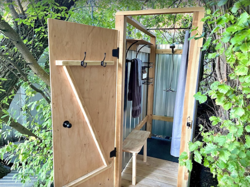 A wooden outdoor shower with an open door, bench, hooks, and surrounded by greenery.