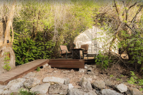A cozy outdoor seating area with a table and chairs near a tent, surrounded by lush greenery and rocky terrain.