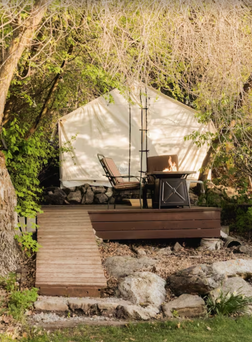 A cozy outdoor setup featuring a tent, a wooden deck, a chair, and a fire pit surrounded by greenery.