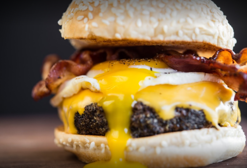 Close-up of a juicy burger with melted cheese, crispy bacon, and a runny egg, all in a sesame seed bun.