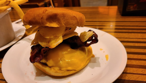 A cheeseburger topped with bacon and potato chips, served on a plate with fries in the background.