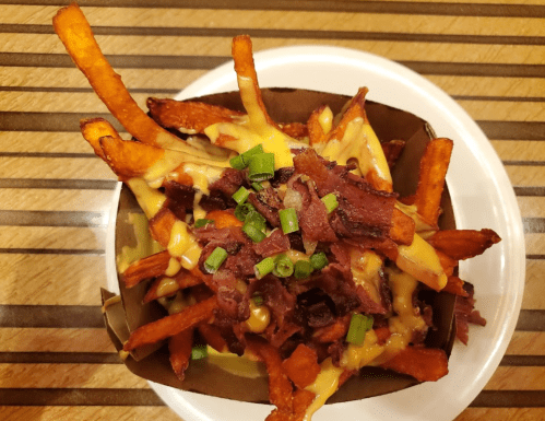 A bowl of loaded sweet potato fries topped with cheese, bacon bits, and green onions on a wooden table.