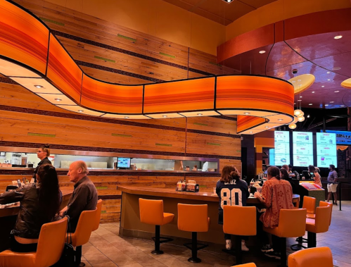 A modern restaurant interior with orange accents, featuring patrons at a curved bar and a menu display in the background.