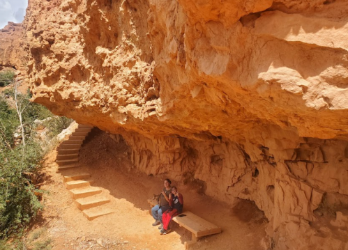 Two people sit on a bench under a rocky overhang, with stairs leading up in a desert landscape.