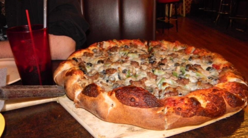 A large pizza topped with sausage, olives, and green peppers, resting on a wooden board next to a drink.
