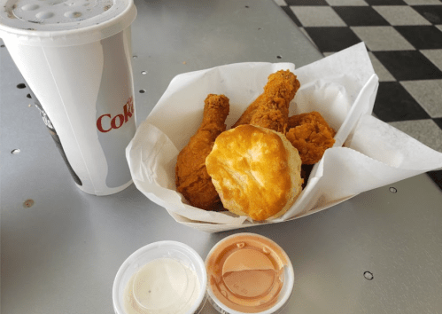 A meal featuring fried chicken legs, a biscuit, dipping sauces, and a cup of soda on a checkered table.