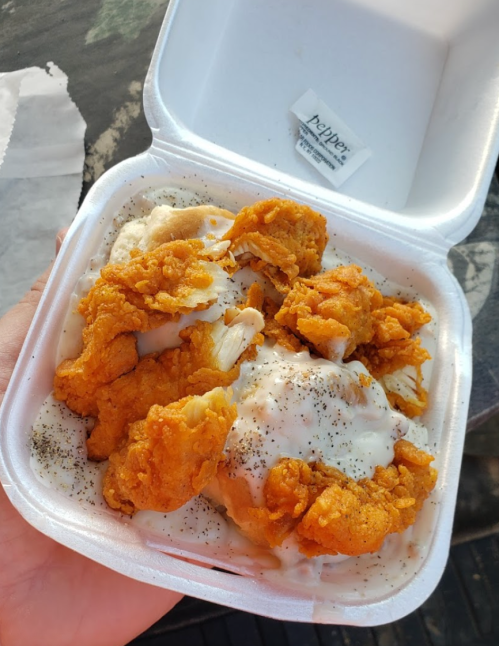 A takeout container filled with fried chicken pieces on top of creamy mashed potatoes and gravy, sprinkled with pepper.