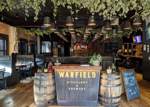 Interior of Warfield Distillery & Brewery featuring wooden barrels, hanging lights, and greenery overhead.