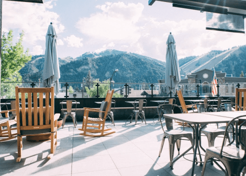 A sunny outdoor patio with wooden rocking chairs and metal tables, overlooking mountains and a clear blue sky.
