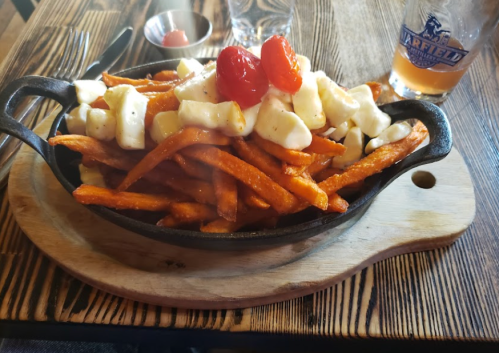 A sizzling skillet of sweet potato fries topped with cheese curds and cherry tomatoes, served on a wooden board.