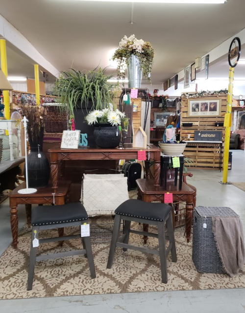 A cozy display of furniture and decor items, including a wooden table, stools, and plants, in a shop setting.