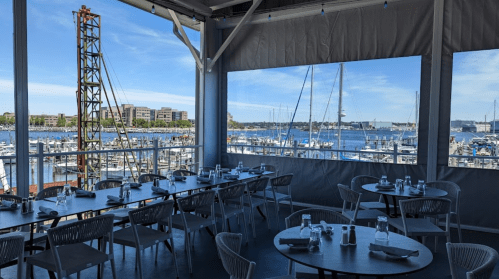 A waterfront restaurant with tables set for dining, overlooking a marina filled with boats and a clear blue sky.