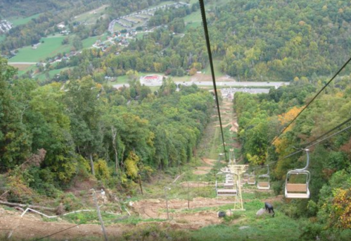 A scenic view from a ski lift overlooking a forested hillside and a valley below, with a road and buildings in the distance.