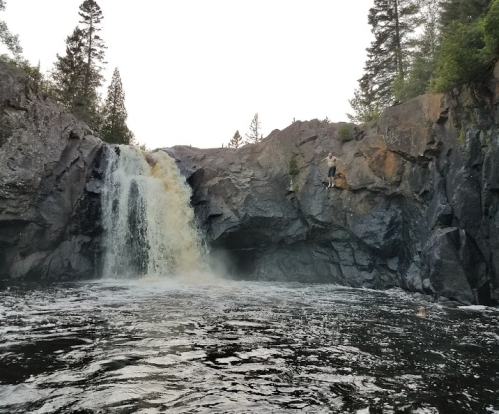 A waterfall cascades over rocky cliffs into a dark pool, surrounded by trees and rugged terrain.