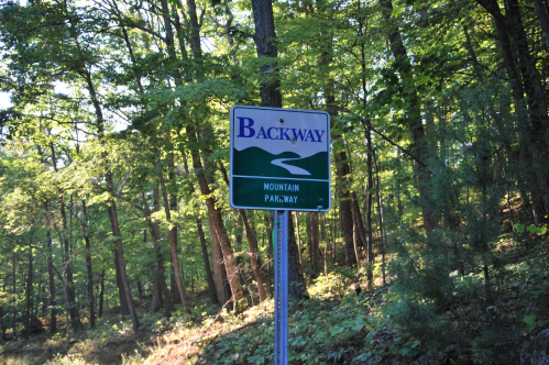 Sign for "Backway Mountain Parkway" surrounded by lush green trees in a forested area.