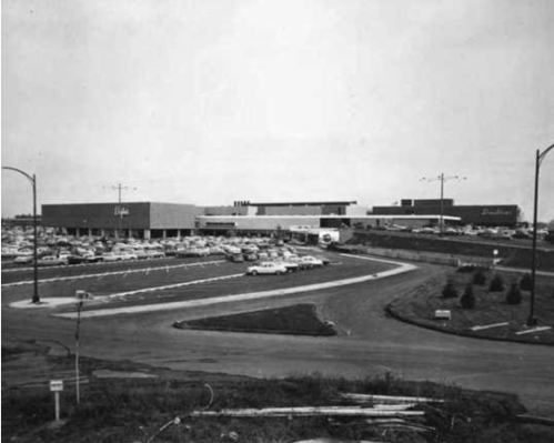 Black and white photo of a shopping center with a large parking lot and surrounding landscape.