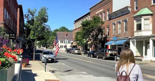 A sunny street scene with shops, parked cars, and an American flag, featuring a person walking away.